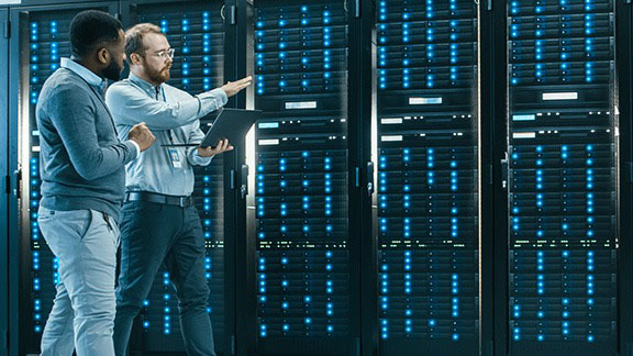 Two men with laptops are talking over a server wall with lots of blue lights.