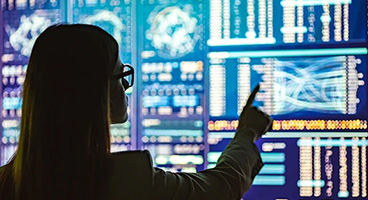 A woman with glasses infront of many screens displaying data