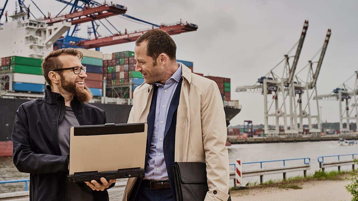 Zwei fröhliche Geschäftsmänner mit Laptop stehen vor großem Hafen mit Containern