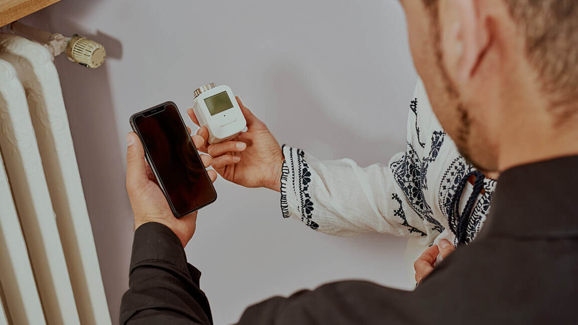 Mann hält Smartphone vor Heizung neben digitalen Thermostat für Heizung