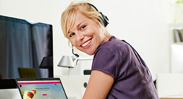 A woman with a headset in front of an open laptop smiles into the camera