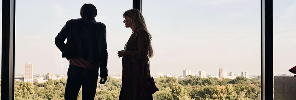 A woman and a man stand in front of a large window and look at the forest and a city beyond.