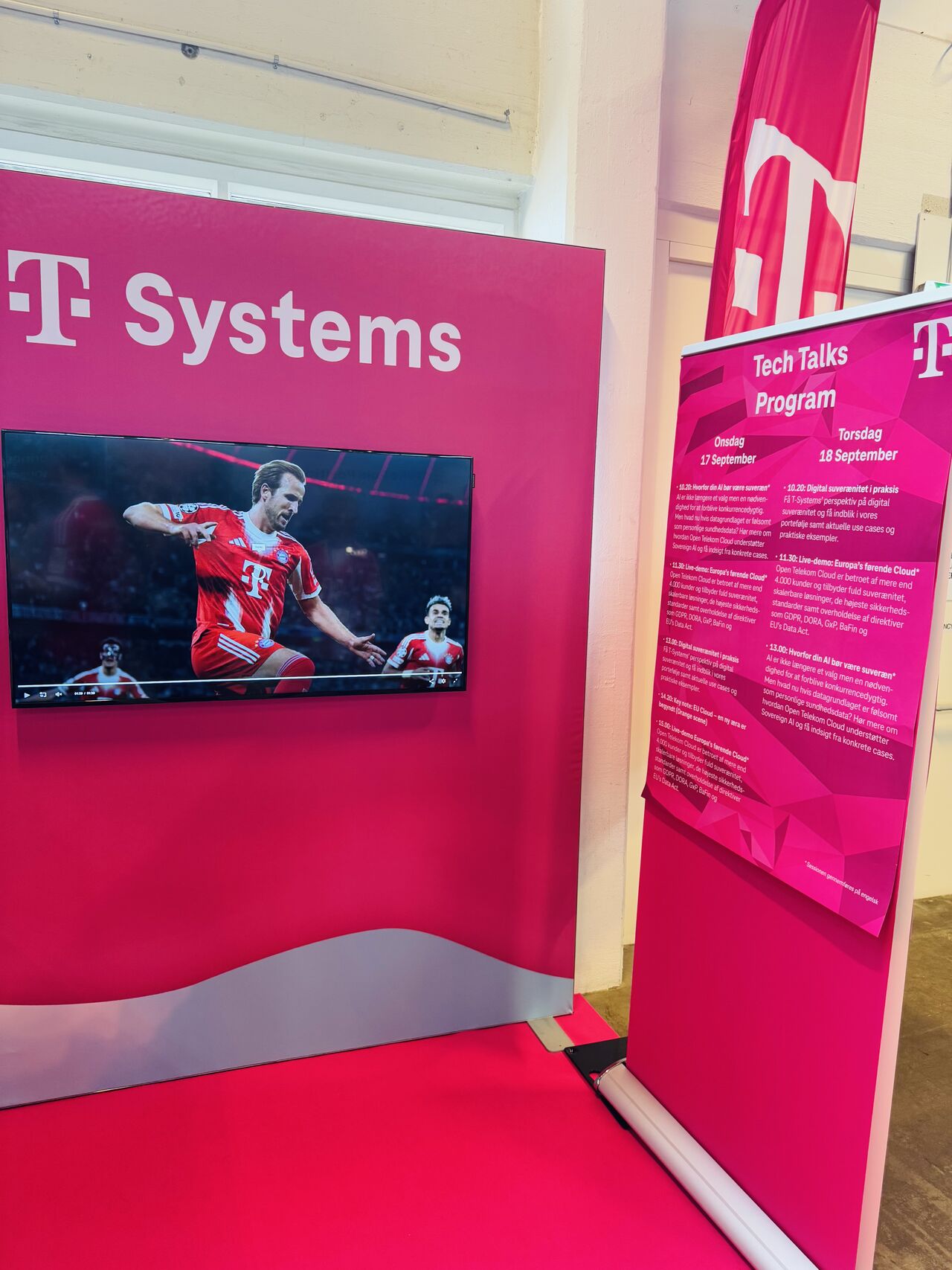 Der Open Telekom Cloud Stand bei mit einem Bildschirm, der ein Fußballspiel zeigt, und einem Veranstaltungshinweis zu den Tech Talks am 17. und 18. September. Der Stand ist in markantem Magenta gehalten.