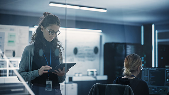 In an office, a woman is standing and looking at a tablet, while another woman is sitting at a PC.