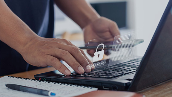 One hand types on a laptop with a security lock hovering above it, the other hand holds a smartphone