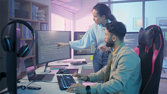 A woman and a man in front of a screen, the woman points to a monitor