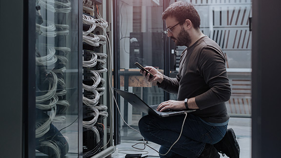 A man with a laptop and phone connected to a server stack.