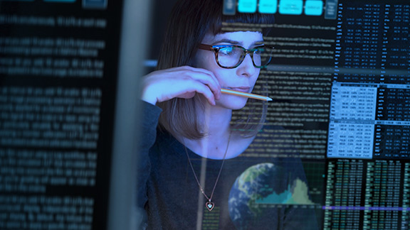 Woman with pen in hand works in front of several screens displaying different data